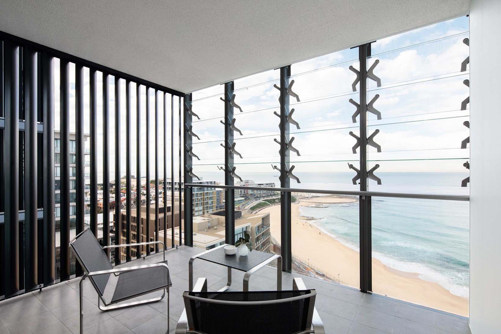 Interior balcony view looking south-east across Newcastle Beach, louvre screens framing the ocean panorama with a chair and table in the foreground
