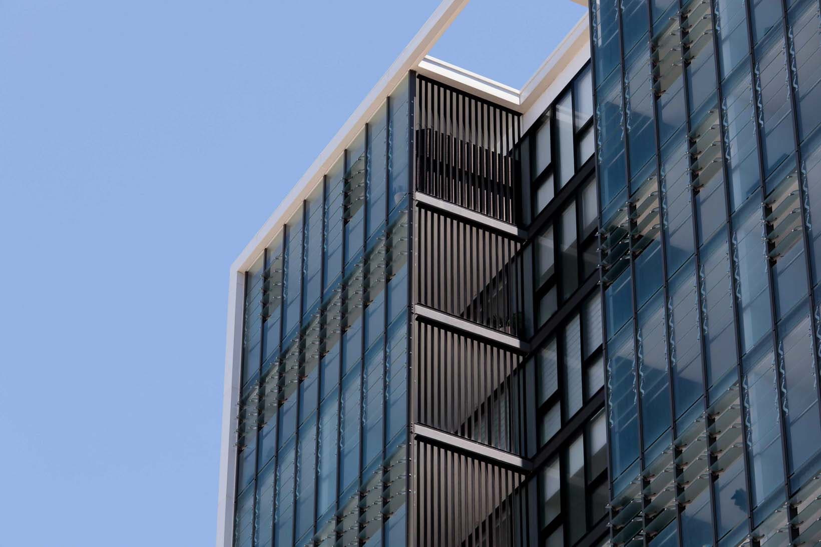 Close-up facade detail, the upper levels of the tower showing the rhythm of glass, vertical louvres and recessed balconies against a clear sky