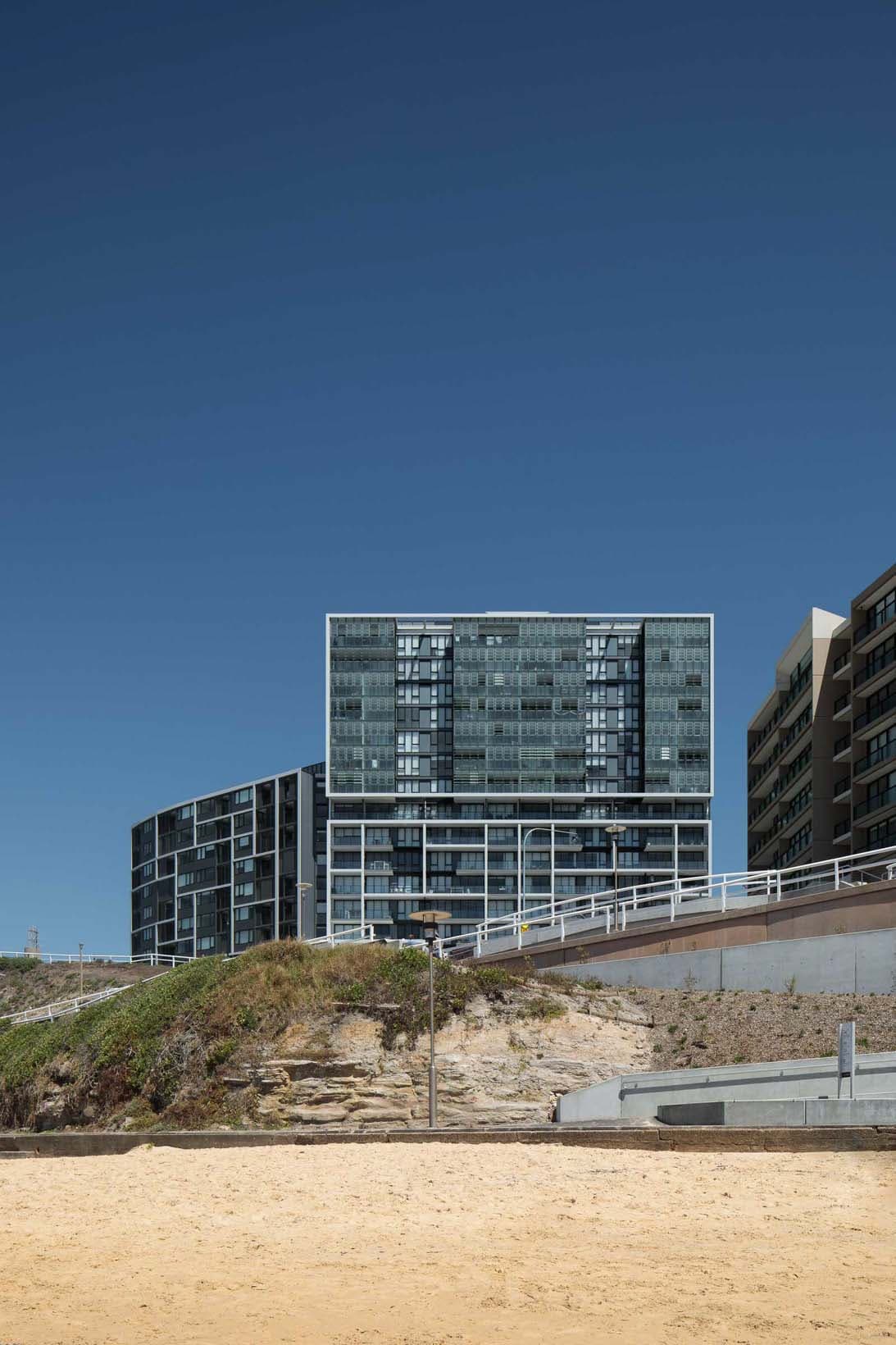 The full Arena development viewed from the beach at dusk, both the curved south building and angular north building visible on the cliff edge above the sandstone escarpment and heritage beach structures
