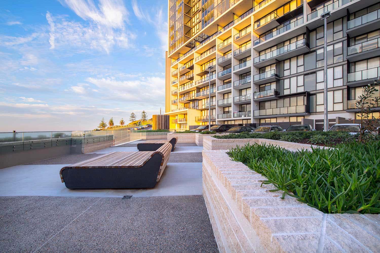 Landscaped podium terrace at golden hour, timber bench seating, planted beds and glass balustrades with the tower rising behind and Norfolk pines visible beyond