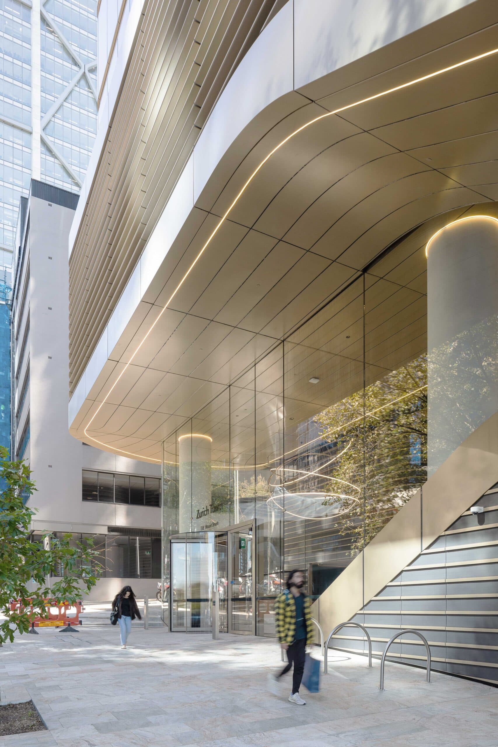 The Zurich Tower entry at ground level, curved gold canopy, glazed lobby and landscaped forecourt with pedestrians