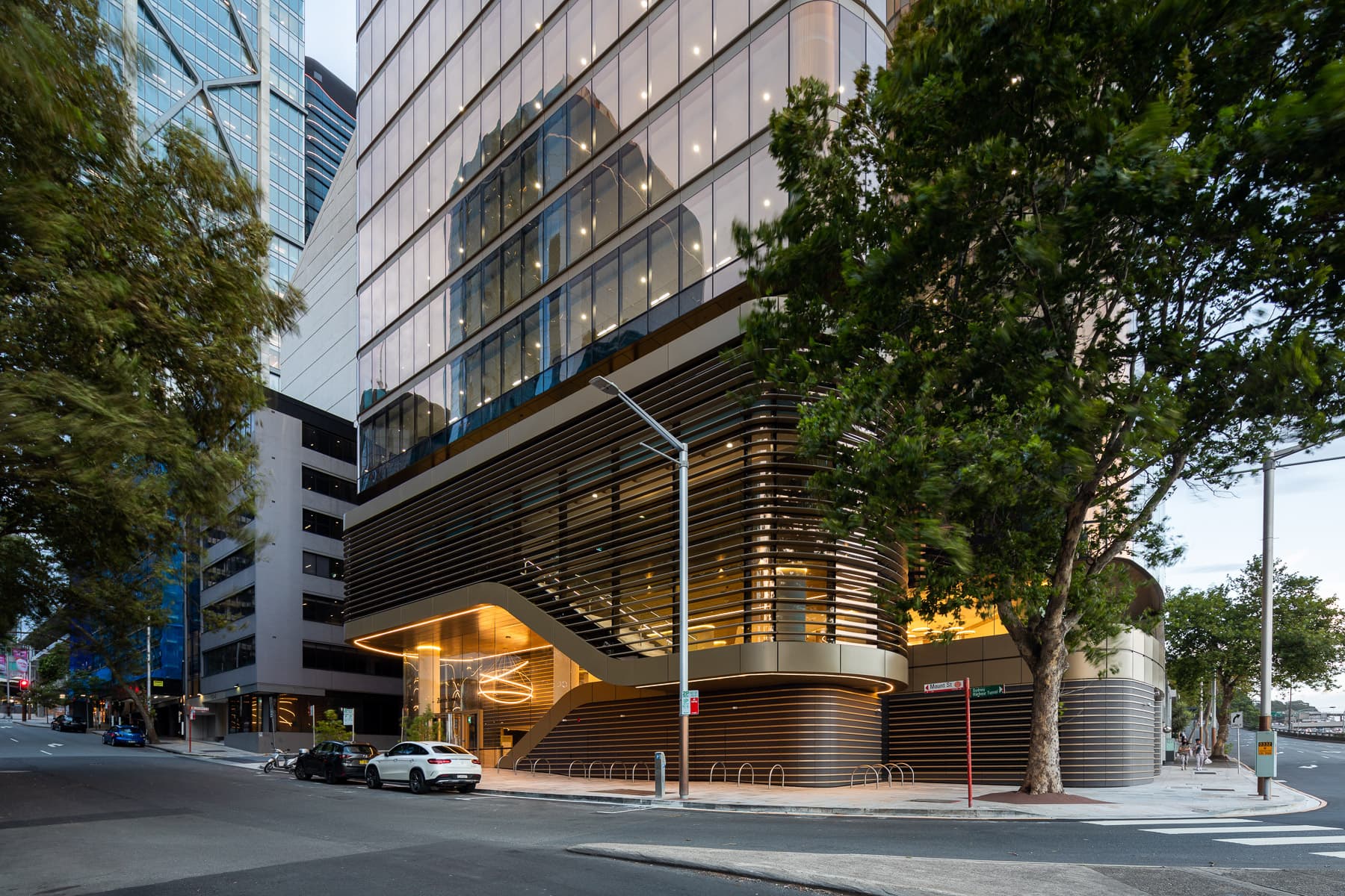 The podium at dusk from the street corner, horizontal bronze louvres wrapping the lower levels with warm light spilling from the lobby behind